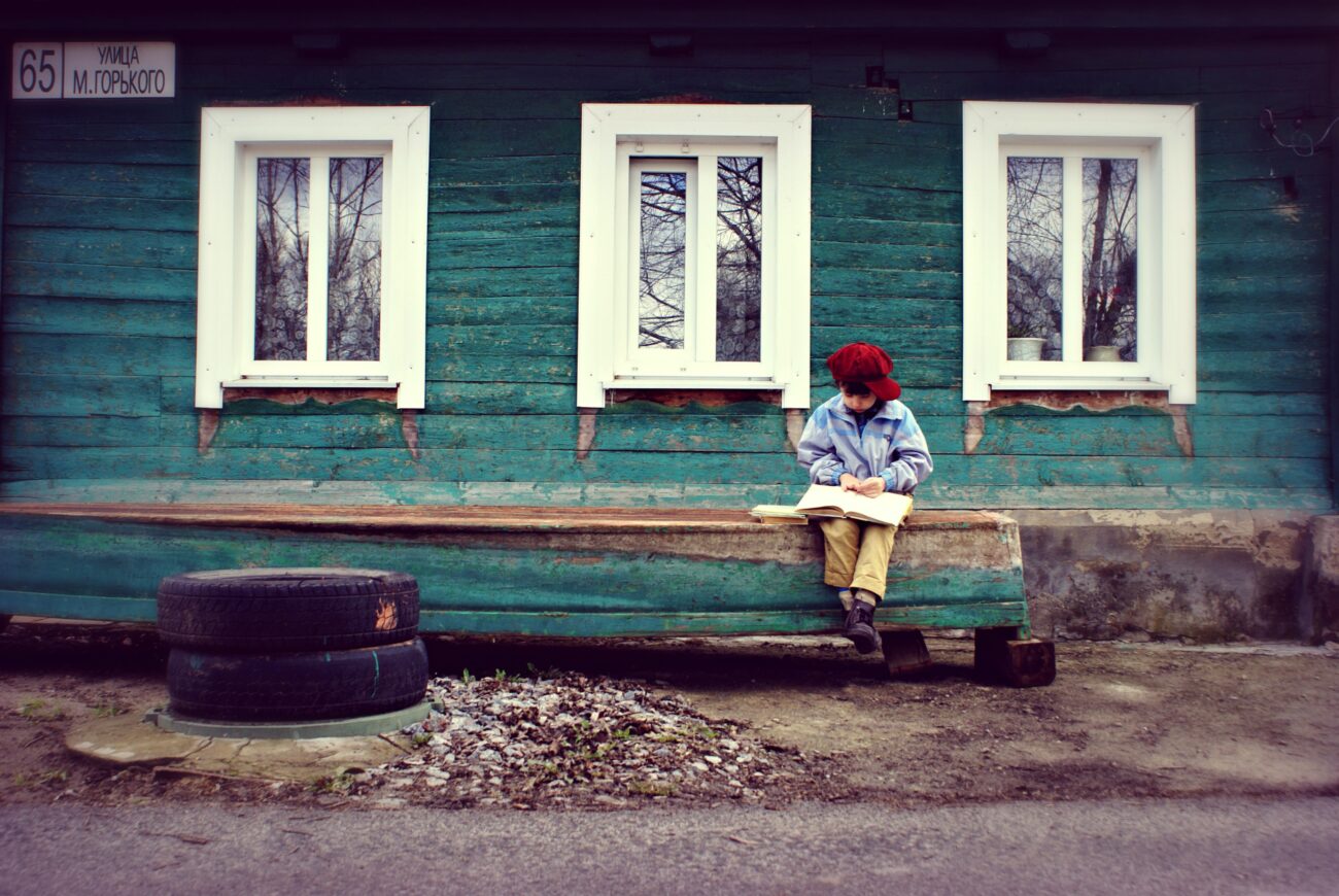 Chłopiec siedzi na drewnianej ławce przed drewnianym domem i czyta książkę.
