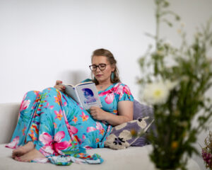 Kobieta w okularach, sukience w kwiaty, siedzi na kanapie i czyta książkę